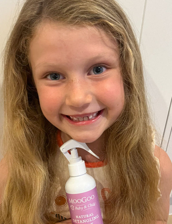 Smiling child with long wavy blonde hair holding a pink-labelled MooGoo detangling spray bottle with a white trigger nozzle.