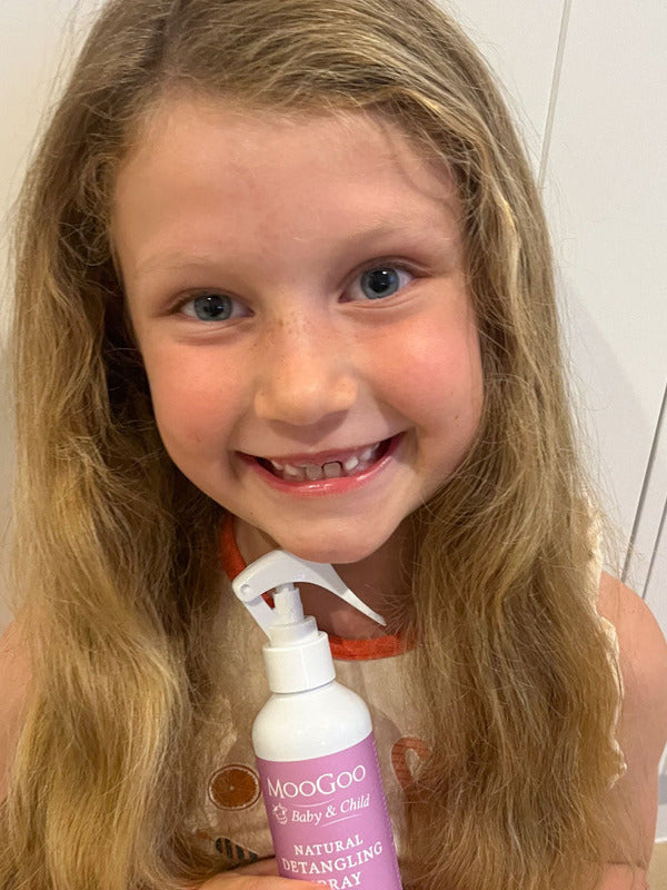 Smiling child with long wavy blonde hair holding a pink-labelled MooGoo detangling spray bottle with a white trigger nozzle.