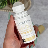 Hand with mint-green nails holding an open MooGoo Oats and Honey deodorant bottle, with its translucent roller ball exposed and tan cap lying behind.