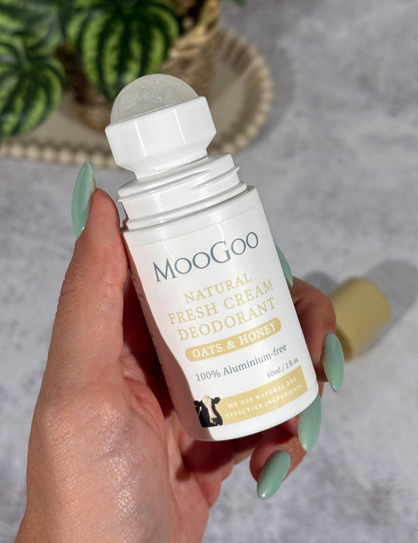 Hand with mint-green nails holding an open MooGoo Oats and Honey deodorant bottle, with its translucent roller ball exposed and tan cap lying behind.