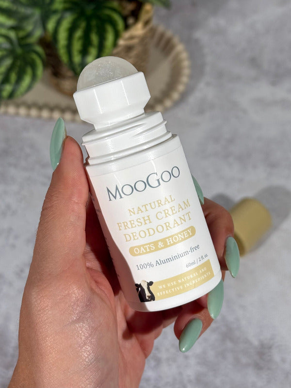 Hand with mint-green nails holding an open MooGoo Oats and Honey deodorant bottle, with its translucent roller ball exposed and tan cap lying behind.