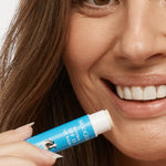 Close-up of a smiling person holding an uncapped blue MooGoo SPF 15 lip balm tube beside their lower lip.