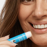 Close-up of a smiling person holding an uncapped blue MooGoo SPF 15 lip balm tube beside their lower lip.