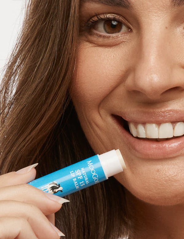 Close-up of a smiling person holding an uncapped blue MooGoo SPF 15 lip balm tube beside their lower lip.