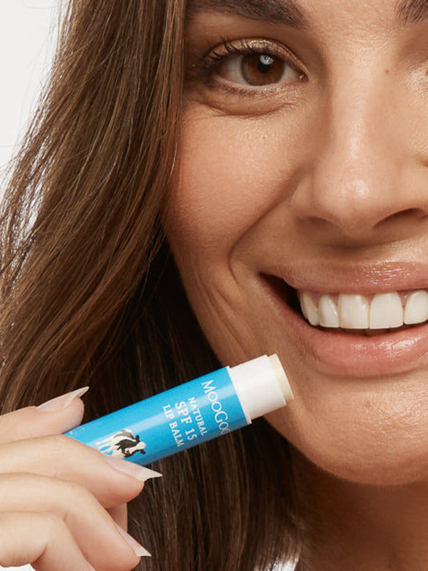 Close-up of a smiling person holding an uncapped blue MooGoo SPF 15 lip balm tube beside their lower lip.