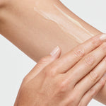 Close-up of a hand spreading a thin white streak of tanning cream over the upper arm against a white background.