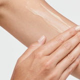 Close-up of a hand spreading a thin white streak of tanning cream over the upper arm against a white background.