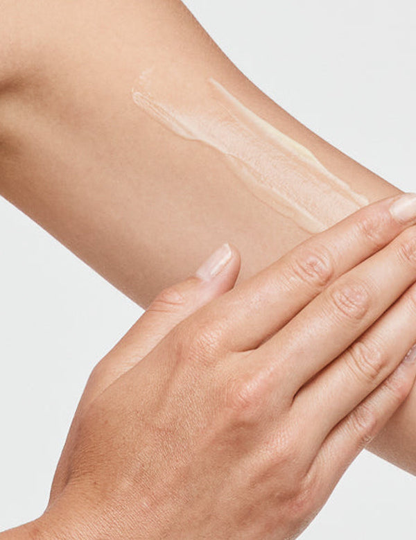 Close-up of a hand spreading a thin white streak of tanning cream over the upper arm against a white background.
