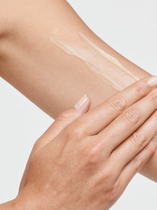 Close-up of a hand spreading a thin white streak of tanning cream over the upper arm against a white background.