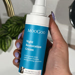 Hand with lilac nail polish holding a blue-labelled MooGoo Night Restoration Cream pump bottle beside a white basin, brass tap and leafy plant.