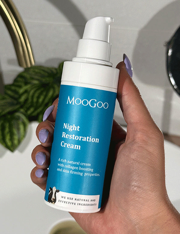 Hand with lilac nail polish holding a blue-labelled MooGoo Night Restoration Cream pump bottle beside a white basin, brass tap and leafy plant.