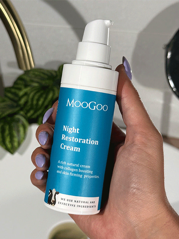 Hand with lilac nail polish holding a blue-labelled MooGoo Night Restoration Cream pump bottle beside a white basin, brass tap and leafy plant.