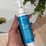 Hand with lilac nail polish holding a tall blue-labelled MooGoo Oil Cleansing Method pump bottle beside a basin on a beige bathroom counter.