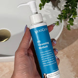 Hand with lilac nail polish holding a tall blue-labelled MooGoo Oil Cleansing Method pump bottle beside a basin on a beige bathroom counter.