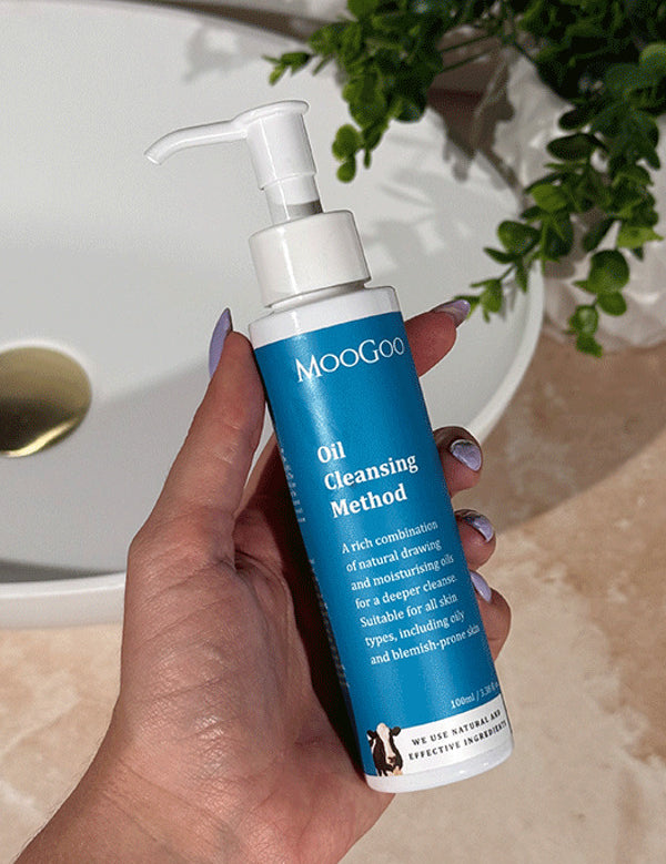 Hand with lilac nail polish holding a tall blue-labelled MooGoo Oil Cleansing Method pump bottle beside a basin on a beige bathroom counter.
