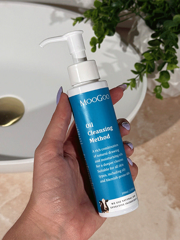 Hand with lilac nail polish holding a tall blue-labelled MooGoo Oil Cleansing Method pump bottle beside a basin on a beige bathroom counter.
