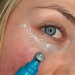 Close-up lifestyle photograph of a woman applying MooGoo 3-in-1 Peptide Eye Serum beneath her eye with the cooling metal roller applicator.