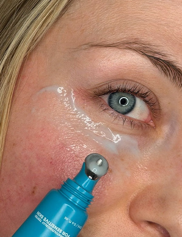 Close-up lifestyle photograph of a woman applying MooGoo 3-in-1 Peptide Eye Serum beneath her eye with the cooling metal roller applicator.