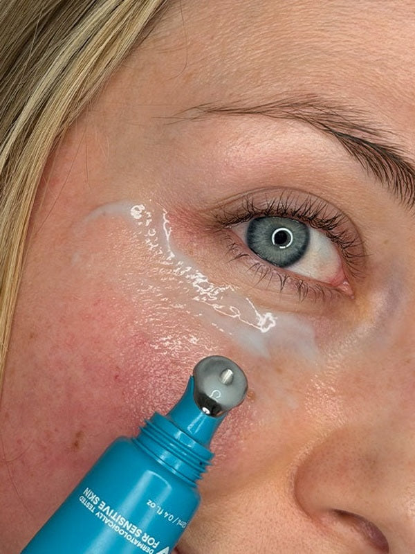 Close-up lifestyle photograph of a woman applying MooGoo 3-in-1 Peptide Eye Serum beneath her eye with the cooling metal roller applicator.