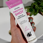 Hand with mint-green nails holding an open white-and-pink MooGoo Protein Shot Leave-In Conditioner tube beside a bathroom basin.