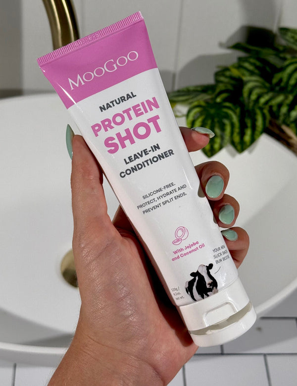 Hand with mint-green nails holding an open white-and-pink MooGoo Protein Shot Leave-In Conditioner tube beside a bathroom basin.