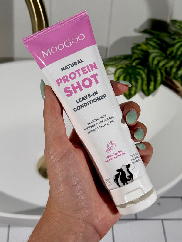 Hand with mint-green nails holding an open white-and-pink MooGoo Protein Shot Leave-In Conditioner tube beside a bathroom basin.