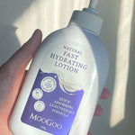 Hand holding a broad white MooGoo Fast Hydrating Lotion pump bottle with a purple label in a bright shaft of sunlight.