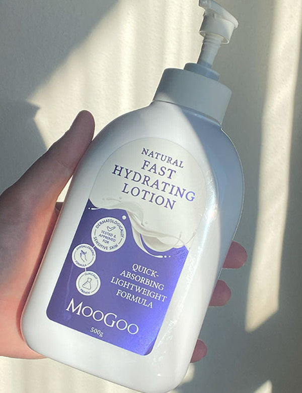 Hand holding a broad white MooGoo Fast Hydrating Lotion pump bottle with a purple label in a bright shaft of sunlight.