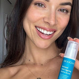 Smiling person holding a blue-labelled MooGoo Retinol Anti-Ageing Active Serum bottle upright beside their face, with its clear cap still on.
