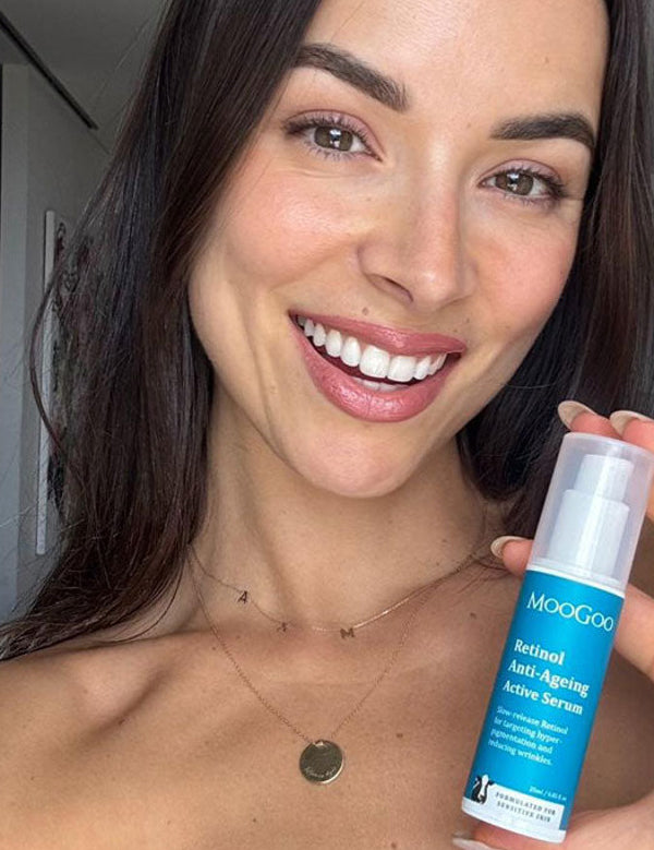 Smiling person holding a blue-labelled MooGoo Retinol Anti-Ageing Active Serum bottle upright beside their face, with its clear cap still on.