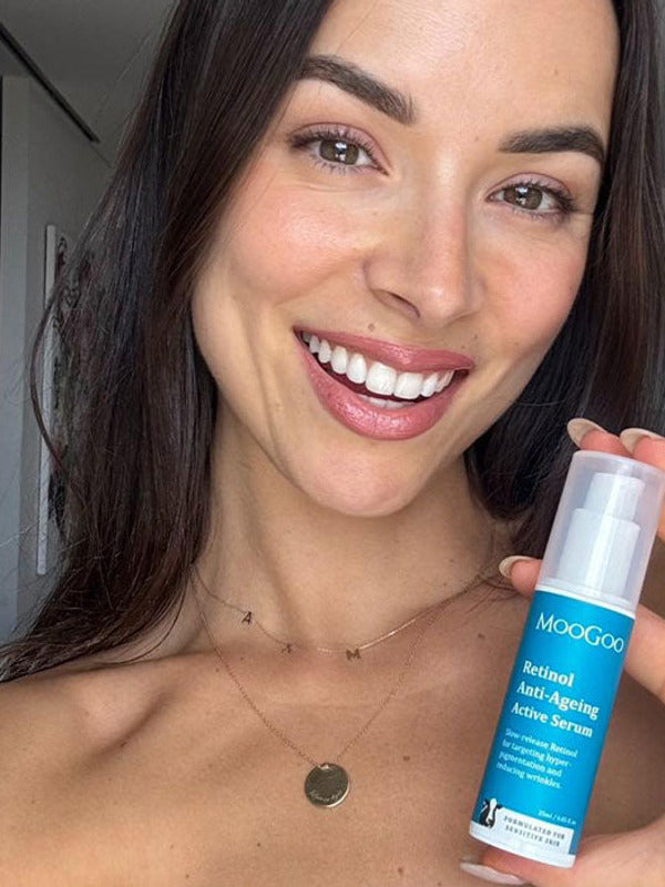 Smiling person holding a blue-labelled MooGoo Retinol Anti-Ageing Active Serum bottle upright beside their face, with its clear cap still on.