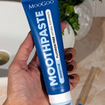 Hand holding a deep blue MooGoo Sensitive Moothpaste tube upright beside a basin, with toothbrushes on the bathroom counter.