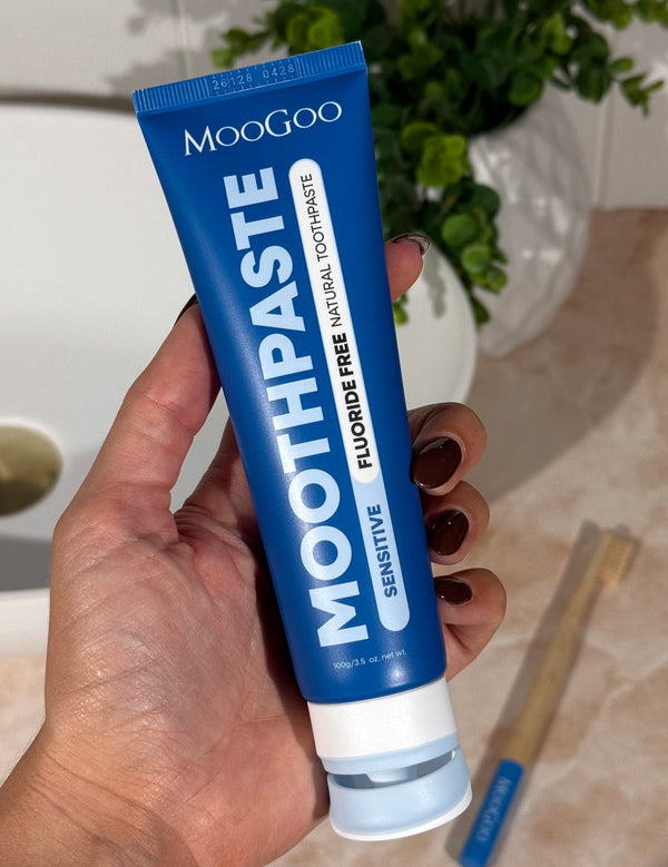 Hand holding a deep blue MooGoo Sensitive Moothpaste tube upright beside a basin, with toothbrushes on the bathroom counter.