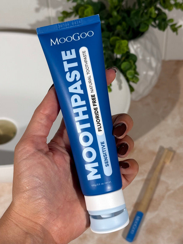 Hand holding a deep blue MooGoo Sensitive Moothpaste tube upright beside a basin, with toothbrushes on the bathroom counter.