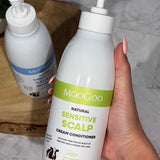 Hand holding a MooGoo Sensitive Scalp Cream Conditioner pump bottle with the matching shampoo on a wooden tray behind.