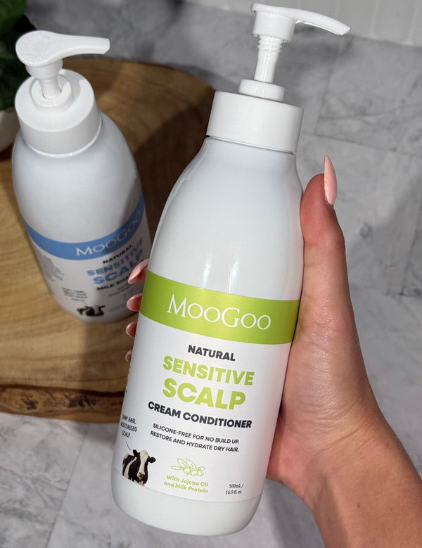 Hand holding a MooGoo Sensitive Scalp Cream Conditioner pump bottle with the matching shampoo on a wooden tray behind.