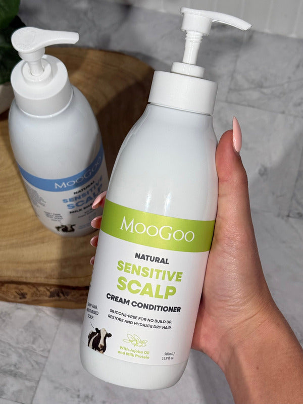 Hand holding a MooGoo Sensitive Scalp Cream Conditioner pump bottle with the matching shampoo on a wooden tray behind.