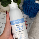 Hand holding a MooGoo Sensitive Scalp Milk Shampoo pump bottle beside a white towel and blue shower puff.