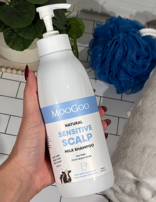 Hand holding a MooGoo Sensitive Scalp Milk Shampoo pump bottle beside a white towel and blue shower puff.