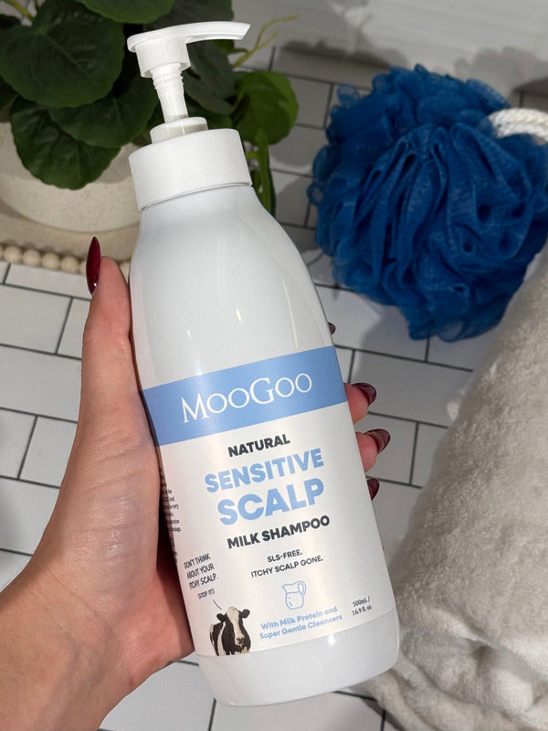 Hand holding a MooGoo Sensitive Scalp Milk Shampoo pump bottle beside a white towel and blue shower puff.