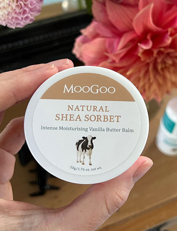 Hand holding a white MooGoo Shea Sorbet tub with its tan lid label facing the camera, in front of pink flowers on a dressing table.
