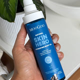 Hand holding an uncapped MooGoo Skin Hero Hydrating Body Oil pump bottle beside a white basin and green leaves.