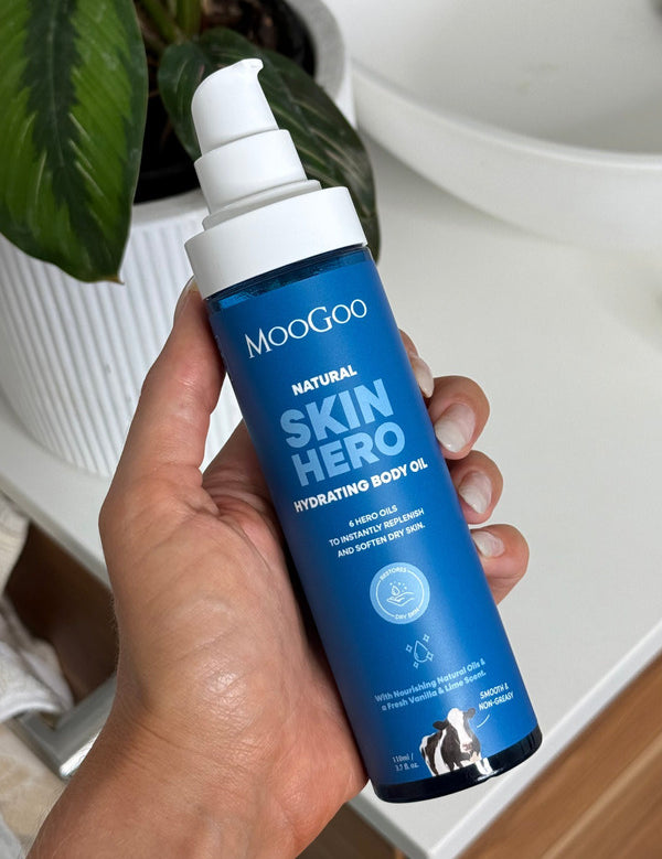 Hand holding an uncapped MooGoo Skin Hero Hydrating Body Oil pump bottle beside a white basin and green leaves.