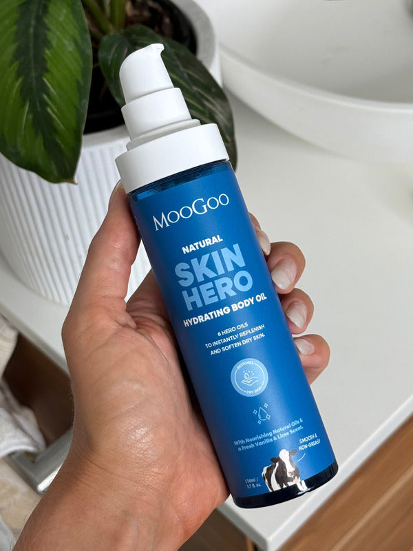 Hand holding an uncapped MooGoo Skin Hero Hydrating Body Oil pump bottle beside a white basin and green leaves.