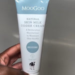 Hand with pale pink nails holding a white-and-pale-blue MooGoo Skin Milk Udder Cream tube with its front label facing the camera.