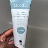 Hand with pale pink nails holding a white-and-pale-blue MooGoo Skin Milk Udder Cream tube with its front label facing the camera.