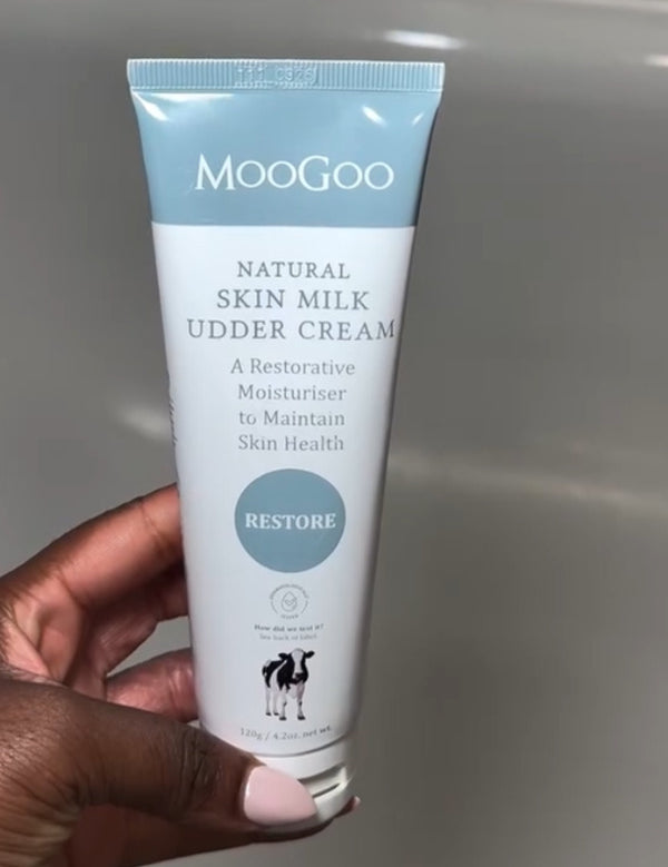 Hand with pale pink nails holding a white-and-pale-blue MooGoo Skin Milk Udder Cream tube with its front label facing the camera.