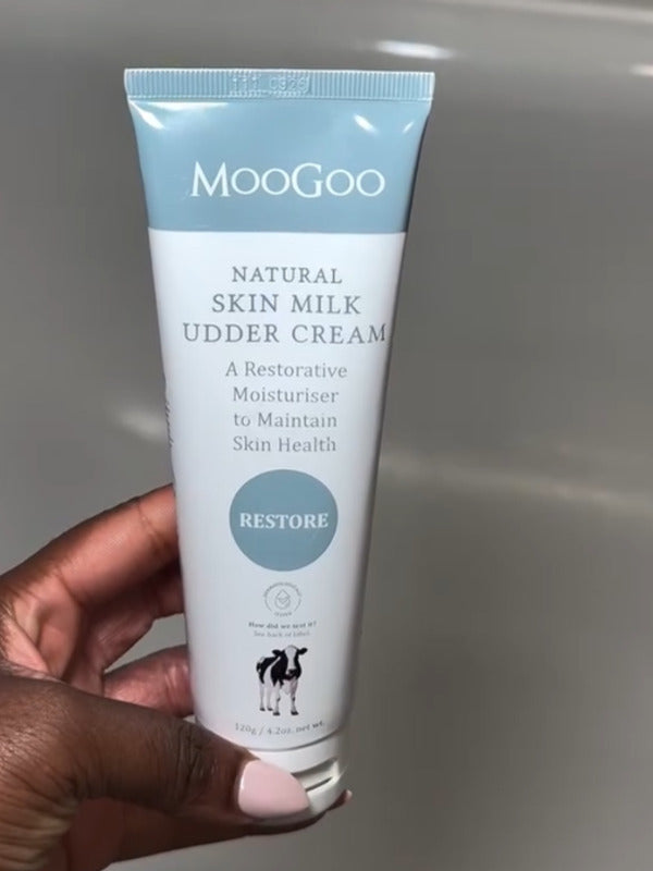 Hand with pale pink nails holding a white-and-pale-blue MooGoo Skin Milk Udder Cream tube with its front label facing the camera.