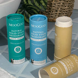 Three MooGoo solid deodorant tubes on blue bathroom tiles: turquoise Lemon Myrtle, blue Coconut Cream and tan Oats and Honey, with the tan stick open and its cap lying in front.