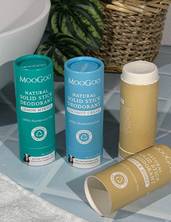 Three MooGoo solid deodorant tubes on blue bathroom tiles: turquoise Lemon Myrtle, blue Coconut Cream and tan Oats and Honey, with the tan stick open and its cap lying in front.